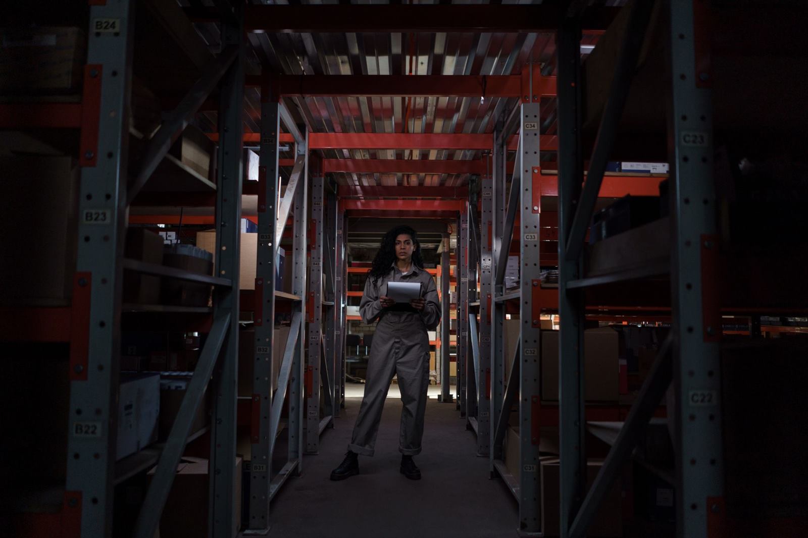 Warehouse worker standing tall among industrial shelves, holding a clipboard in dim lighti